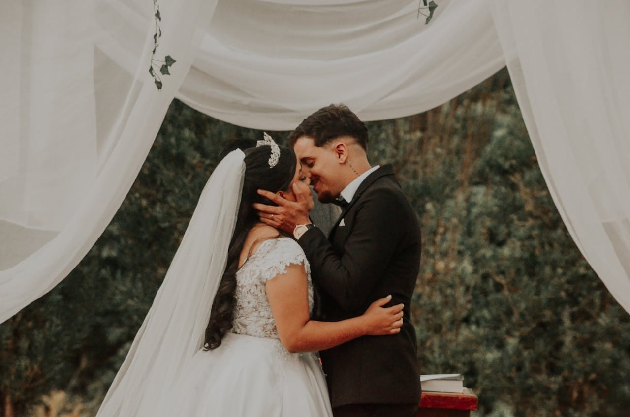 services-img Bride and groom share a romantic kiss during an outdoor wedding ceremony surrounded by elegant drapery.